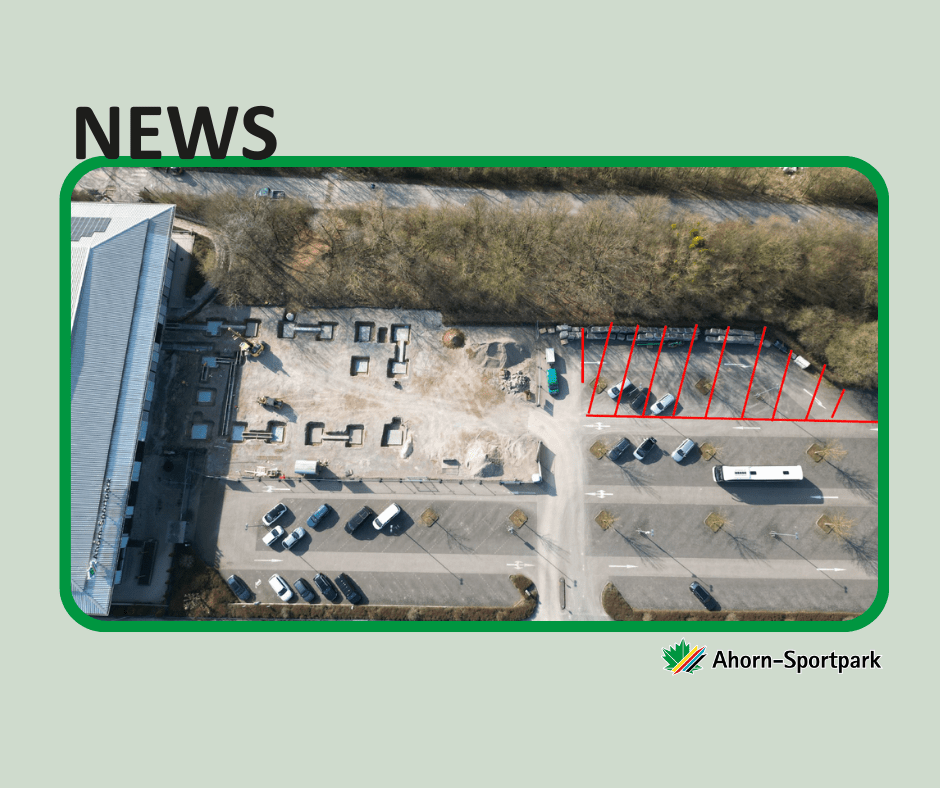 Luftaufnahme des Ahorn-Sportparks mit einer Baustelle neben der Sporthalle. Auf dem Gelände sind Bauarbeiten und Fundamentstrukturen zu sehen. Rechts daneben ist ein Parkplatzbereich, in dem mehrere Parkplätze mit roten Linien markiert und aktuell gesperrt sind. Im Vordergrund befinden sich weitere Parkplätze und eine Zufahrtsstraße.