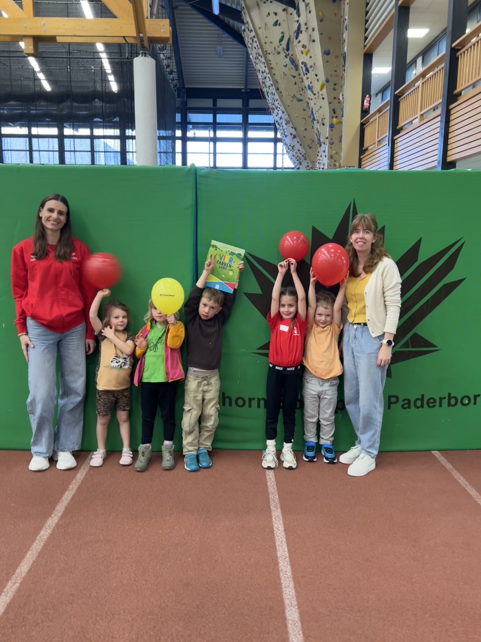 Das Foto zeigt sieben Personen in einer Reihe vor einer grünen Prallwand/Matte aufgestellt. Links und rechts außen stehen zwei Frauen, dazwischen sind fünf Kinder die je einen Luftballon in der Hand hochhalten. Ein Kind hat ein Buch mit dem Titel "Vier-Farben-Land" hoch in den Händen. Alle Personen lachen in die Kamera. Das Foto entstand im Zusammenhang mit dem Aktionstak gegen Rassismus. Dort lernte die Kinder spielerisch etwas über Rassismus und Ausgrenzung