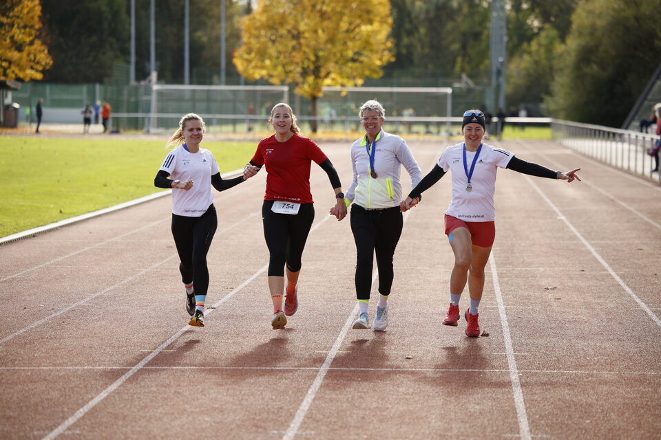 Zu sehen ist der gemeinsame Zieleinlauf von Laura Terheiden, Jutta Savis, Katrin Deutsch und Annika Monkau vom Team LC Stefan (Staffel-Team) beim 8. Almetal-Marathon im Leihtathletikstadion des Ahorn-Sportpark. Im Hintergrund sind die Kleinspielfelder mit einem Baum, welcher herbstliche Farben trägt zu sehen.