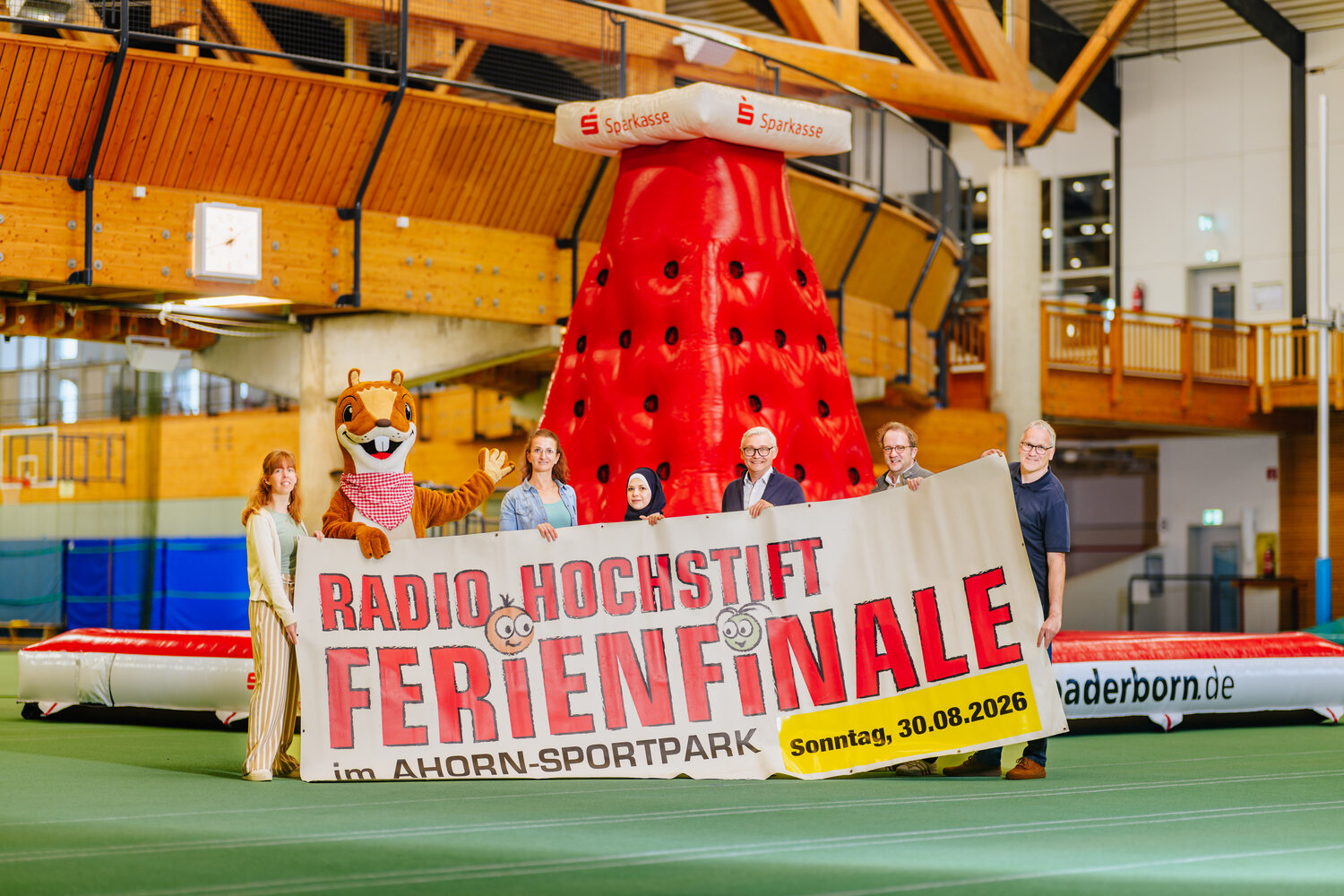 Im Vordergrund stehen die Verantwortlichen des Radio Hochstift-Ferienfinales mit dem Ankündigungsbanner des RHFs vor dem Kletterfelsen. Das Ferienfinale findet am 30.08.2026 statt.