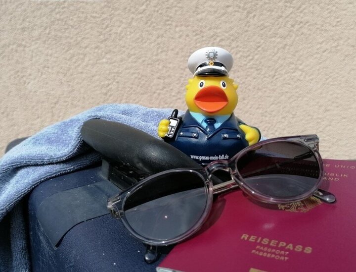 Eine Polizei-Gummiente sitzt auf einem Koffer. Auf dem Koffer liegt vor der Ente eine Sonnenbrille und ein deutscher Reisepass.