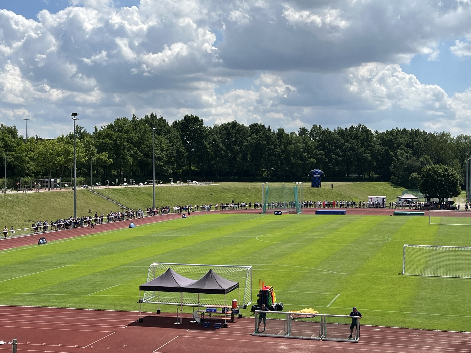 Weitblick über den Ahorn-Sportpark zur Saisoneröffnung des SC Paderborn 07: Auf dem Rasenplatz mit umlaufender Laufbahn haben sich zahlreiche Besucherinnen und Besucher entlang der Strecke versammelt. Im Vordergrund stehen zwei Pavillons am Spielfeldrand, im Hintergrund rahmen Bäume und ein bewölkter Himmel die Sportanlage ein.