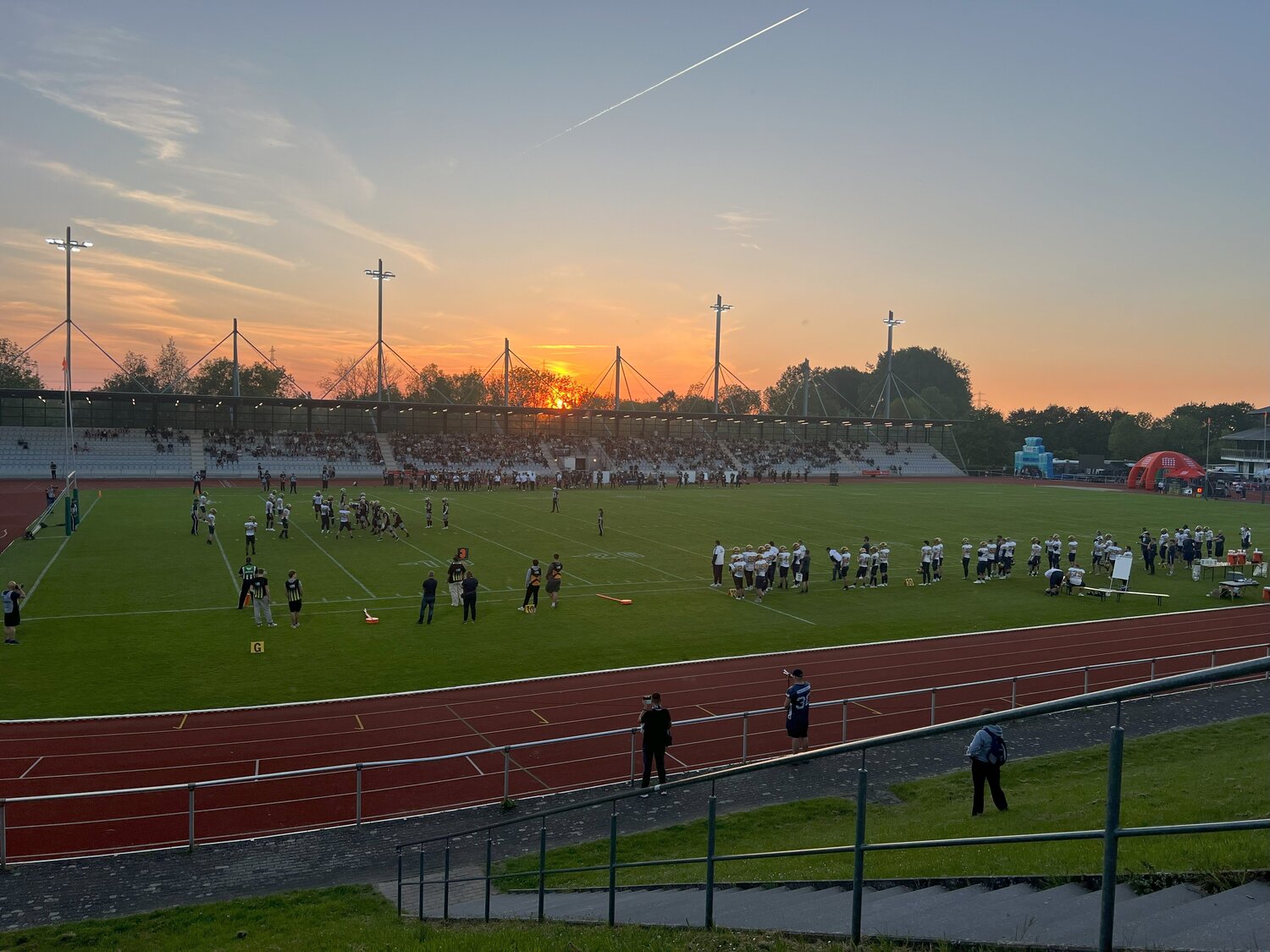 Weitwinkelaufnahme des Ahorn-Sportparks während eines American-Football-Spiels der Paderborn Dolphins. Auf dem Spielfeld stehen beide Teams in Spiel- und Seitenlinienformation, während auf der gegenüberliegenden Tribüne zahlreiche Zuschauer das Spiel verfolgen. Im Hintergrund geht die Sonne hinter der Tribüne unter und taucht das Stadion in warmes Abendlicht.