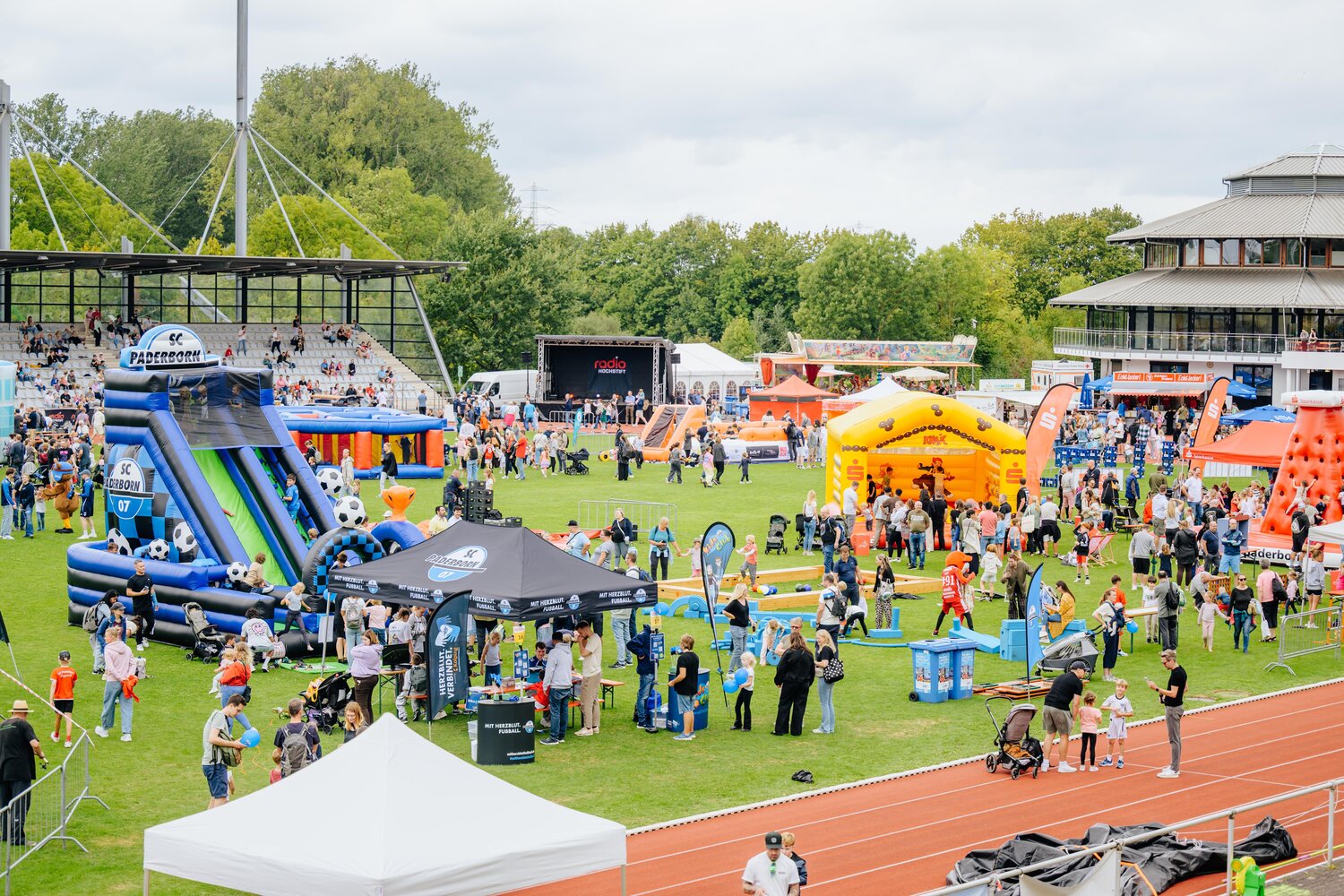 Blick über das gut besuchte Stadion des Ahorn-Sportparks beim Radio Hochstift-Ferienfinale. Auf dem Rasen verteilen sich zahlreiche Familien zwischen bunten Hüpfburgen, Aktionsständen und Mitmachangeboten. Im Hintergrund befindet sich die Bühne mit Showprogramm.
