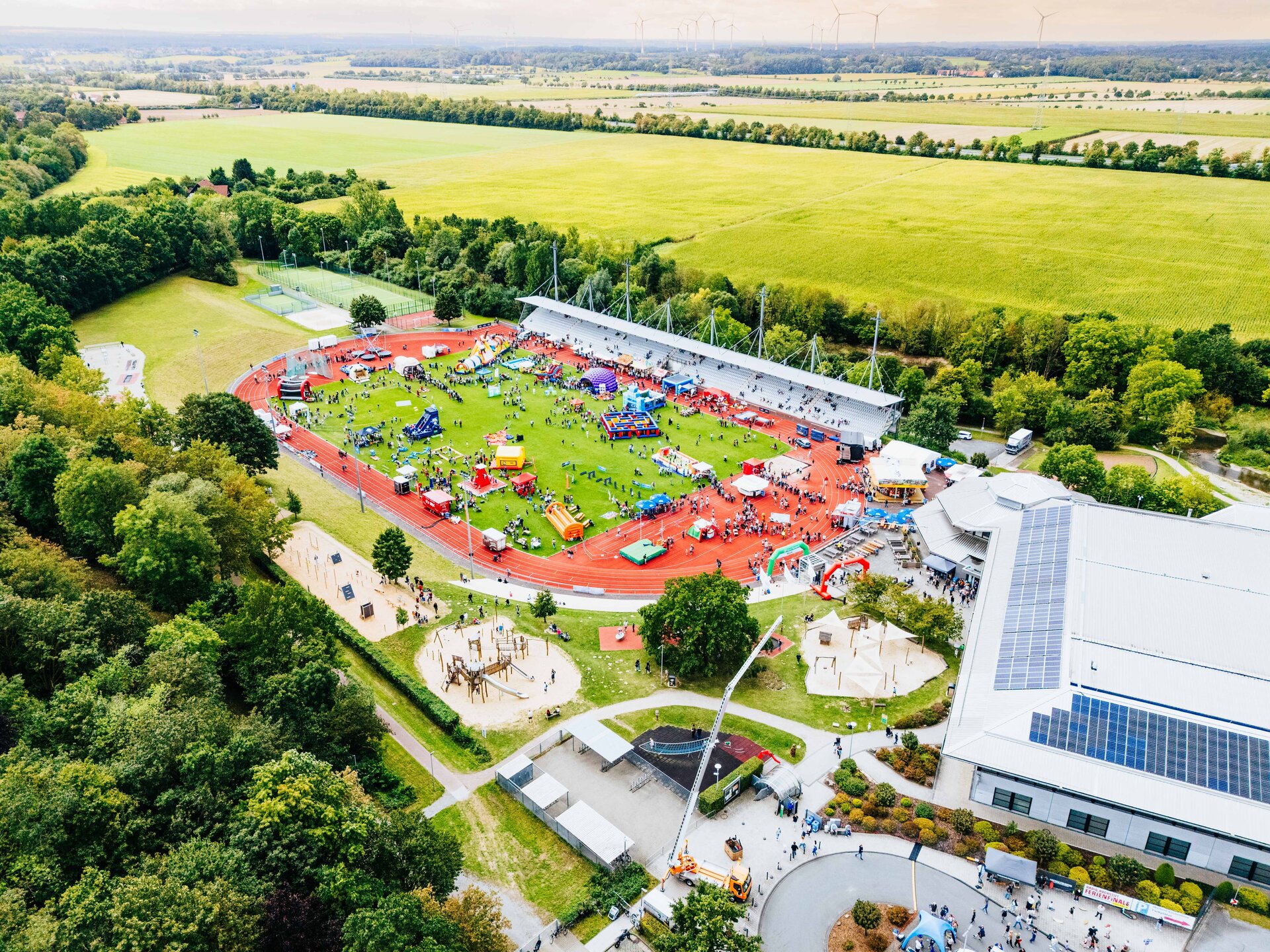 Luftaufnahme des Ahorn-Sportparks während des Radio Hochstift-Ferienfinales. Im Stadion sind zahlreiche Besucherinnen und Besucher zwischen bunten Hüpfburgen, Aktionsständen und Mitmachangeboten unterwegs. Rund um die Sportanlage erstrecken sich grüne Felder und Waldflächen.