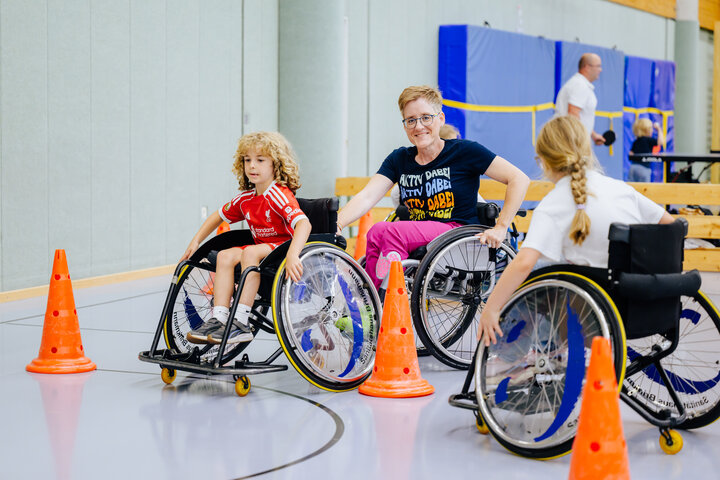 Kinder und eine erwachsene Person absolvieren beim Radio Hochstift-Ferienfinale einen Parcours mit Sportrollstühlen in der Sporthalle des Ahorn-Sportparks. Orangefarbene Hütchen markieren den Parcours.