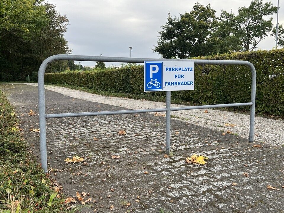 Ein Absperrgitter steht vor einem Weg. An dem Gitter hängt ein Schild auf dem "Parkplatz für Fahrräder steht"