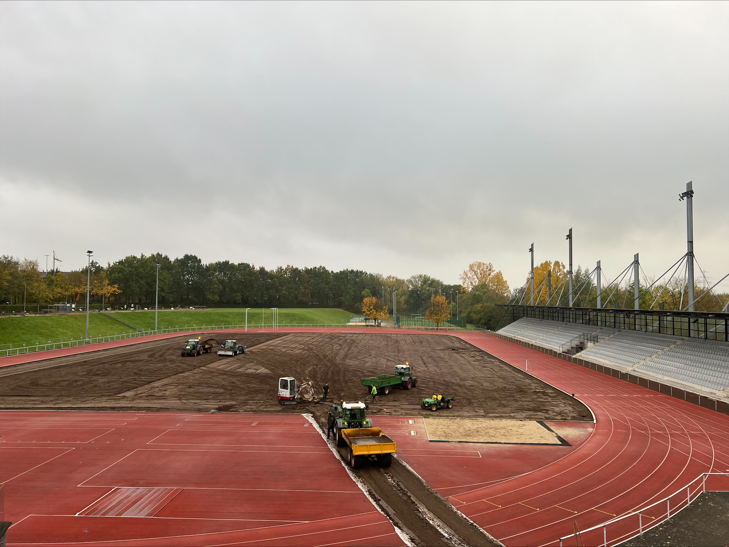 Blick auf das Leichtathletikstadion im Ahorn-Sportpark während der Sanierung des Stadionrasens. Die Rasenfläche wurde bereits vollständig abgetragen. Mehrere Baufahrzeuge und Arbeiter sind auf der freigelegten Erdfläche im Einsatz. Die rote Tartanbahn und die Stadiontribüne sind im Hintergrund und Vordergrund zu sehen.
