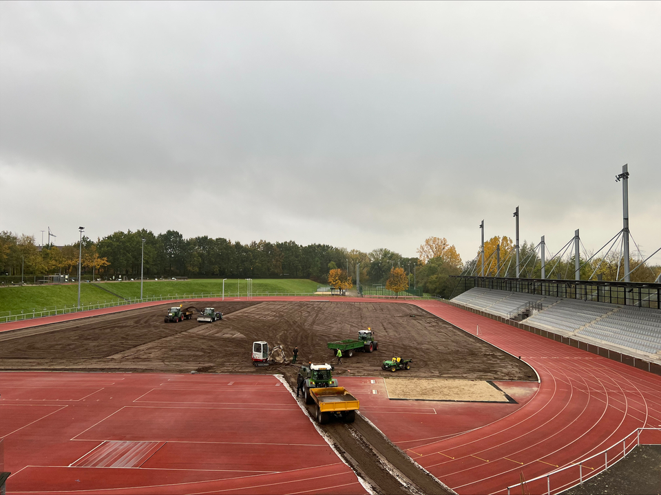 Blick auf das Leichtathletikstadion im Ahorn-Sportpark während der Sanierung des Stadionrasens. Die Rasenfläche wurde bereits vollständig abgetragen. Mehrere Baufahrzeuge und Arbeiter sind auf der freigelegten Erdfläche im Einsatz. Die rote Tartanbahn und die Stadiontribüne sind im Hintergrund und Vordergrund zu sehen.