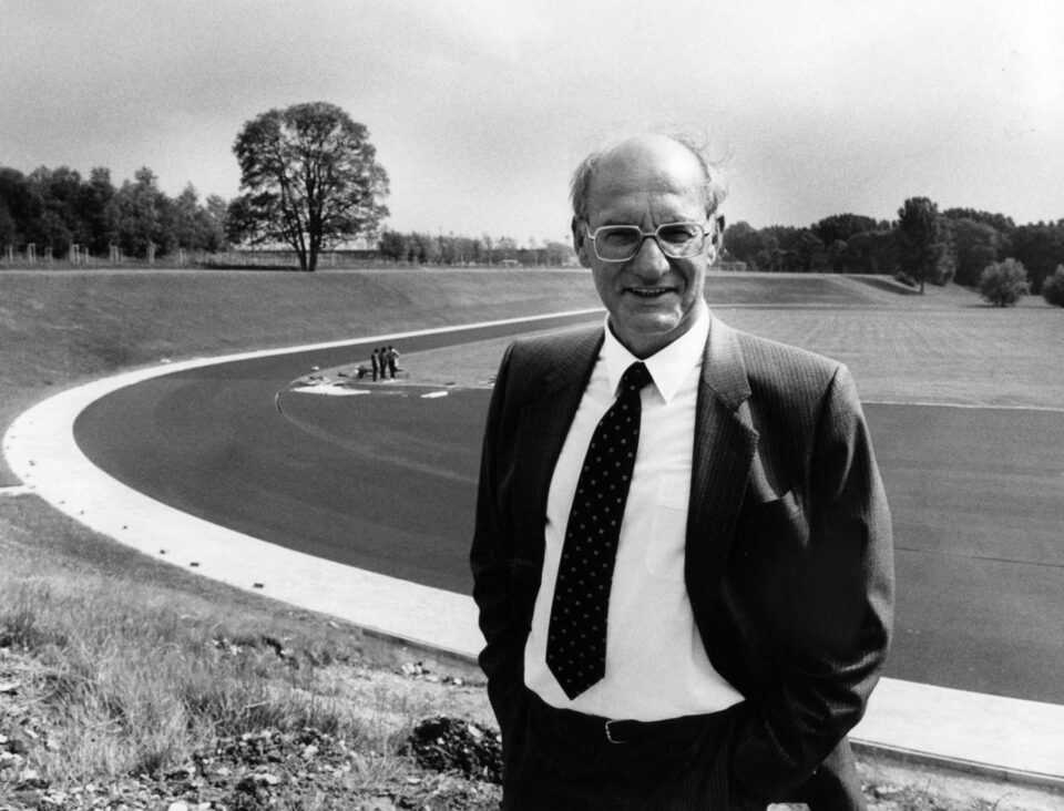 Auf diesem schwaz-weiß Foto von Heinz Nixdorf steht er vor dem Leichtathletikstadion mit Blick nach vorne. Im Hintergrund ist die Leichtathletikfläche mit dem Rasen und der Tartanbahn zu erkennen. Weiter im Hintergund sind Bäume zu sehen.